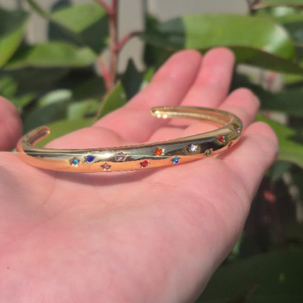 A gold-toned open cuff bracelet with small colorful star-shaped rhinestones, held in a person's hand against a background of green leaves.