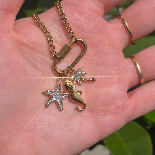 Gold-toned necklace with a lobster clasp and dangling starfish and seahorse charms, shown resting on an open hand. The starfish charm is embellished with small clear stones.