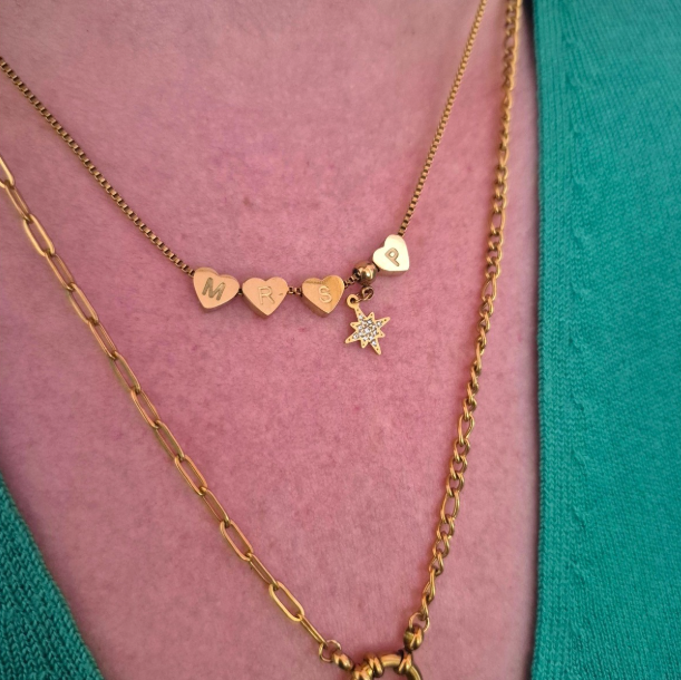 A close-up of a person wearing layered gold necklaces over a pink top and teal cardigan. The necklace features three small heart charms engraved with letters, a small star charm, and a delicate chain.