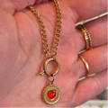 A gold chain necklace with a toggle clasp and a round pendant featuring a red heart, held in a person's hand.