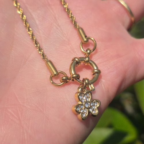 A gold chain necklace with a toggle clasp and a dangling flower charm set with clear stones, resting on a person's hand.