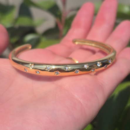 A gold-toned open cuff bracelet with small clear stones set along the front, held in a person's hand against a blurred green outdoor background.